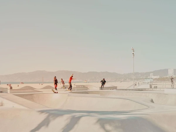 A group of skateboarders and scooter riders perform tricks in a concrete skatepark on a sunny beach in an outdoor recreation scene.
