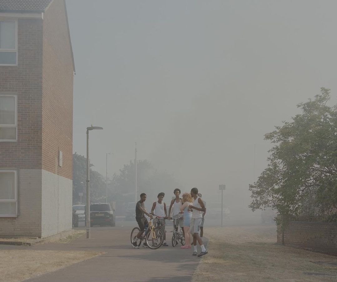 Group of young adults with bikes stand on a path next to a brick building; hazy light; street with cars parked along the sides.