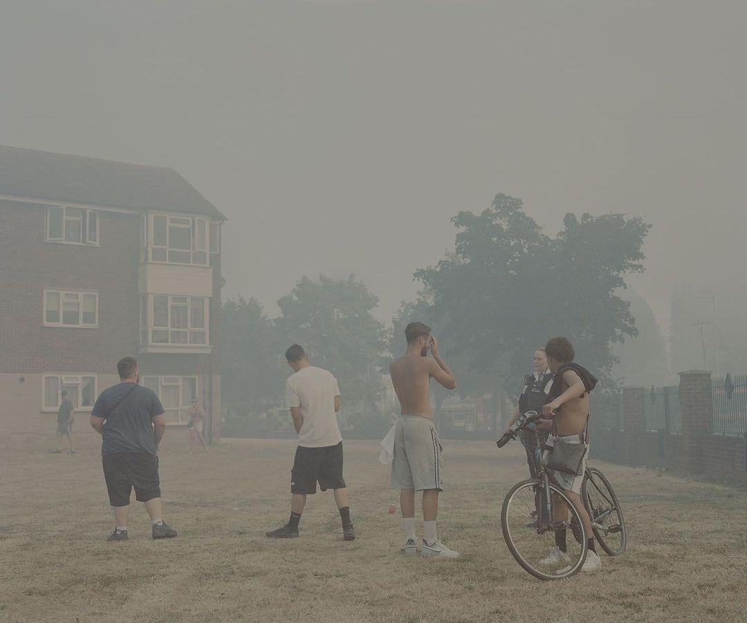 Outdoor scene featuring a group of young adult males with bicycles in a smoky haze near a brick building and trees, on a grassy area.