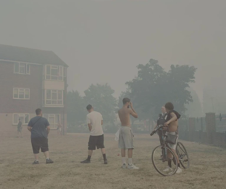 Outdoor scene featuring a group of young adult males with bicycles in a smoky haze near a brick building and trees, on a grassy area.