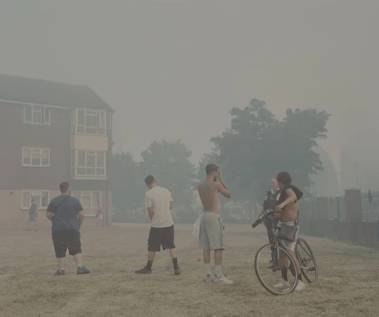 Outdoor scene featuring a group of young adult males with bicycles in a smoky haze near a brick building and trees, on a grassy area.