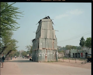 Tall makeshift tower constructed of corrugated metal sheets standing in the middle of a dusty wide street with people