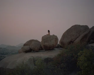 A person sitting in meditation atop a large rounded boulder amid a rugged rocky landscape under an overcast sky.