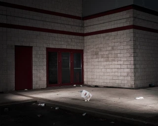 A crumpled plastic bag rests on a paved area in front of a light brick building exterior with dark red doors and trim.