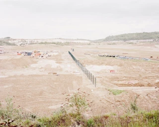 Wide landscape shot of a vast construction site with exposed earth, piles of materials, and a long security fence dividing the terrain.