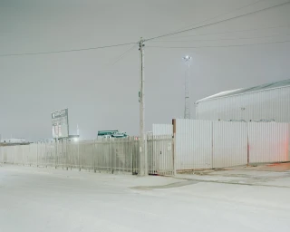 Industrial warehouse building exterior with metal fencing and utility pole under an overcast sky at night
