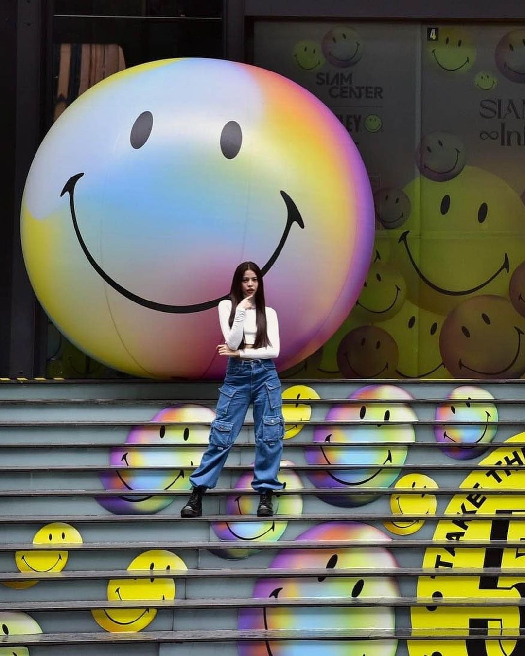 Young woman poses on stairs decorated with smiley faces, next to large colorful smiley face balloon and wall of repeating smiley faces.