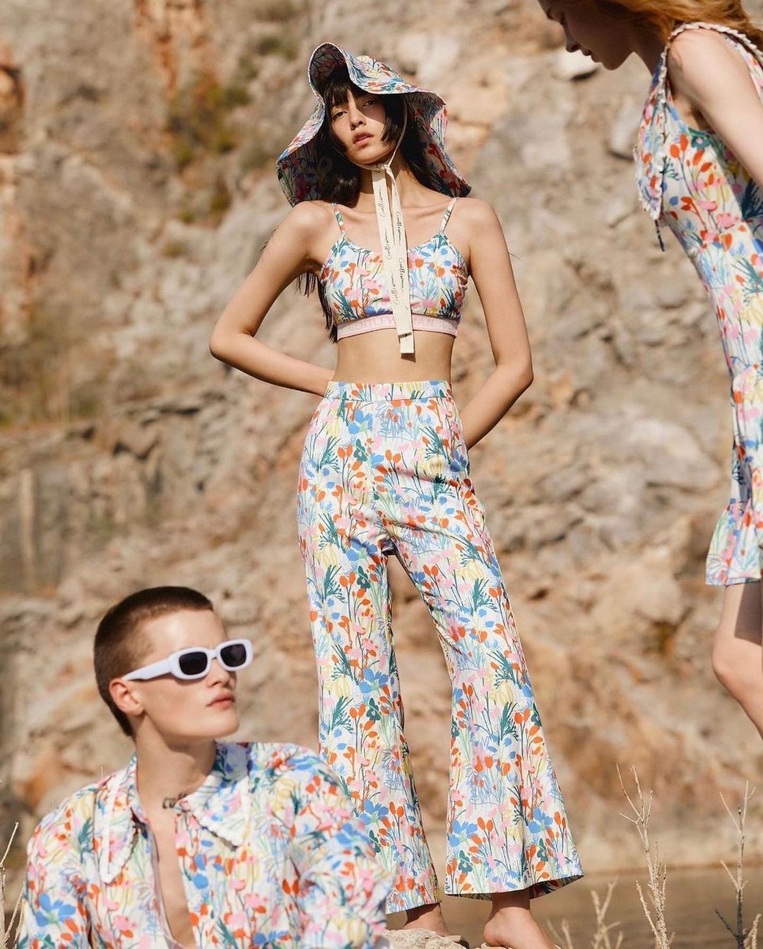 Three models in floral print outfits pose against a rocky background, one seated with white sunglasses, others standing in the daytime.