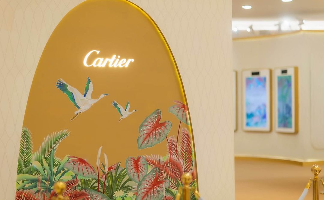 Cartier storefront display with a gold background, two white birds in flight, and tropical plants, creating a luxurious natural scene.