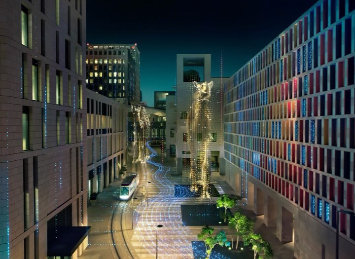 Modern city street view at night with illuminated buildings and sculptures, featuring a tram on the tracks and carefully placed greenery.