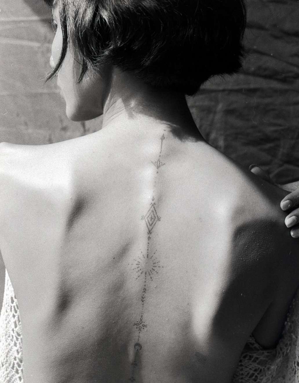 Back view of a person with a geometric tattoo design running down the spine against a textured backdrop with a partial view of a white lace shawl.