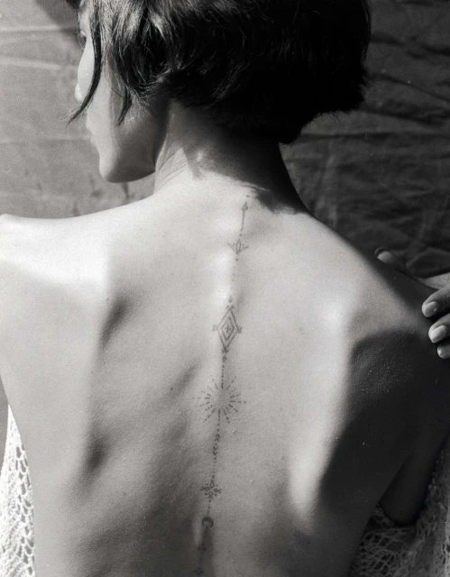 Back view of a person with a geometric tattoo design running down the spine against a textured backdrop with a partial view of a white lace shawl.