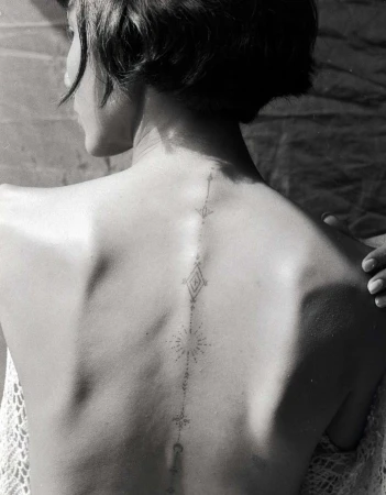 Back view of a person with a geometric tattoo design running down the spine against a textured backdrop with a partial view of a white lace shawl.