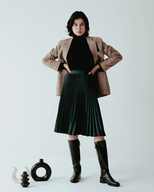 Fashionable woman posing in pleated skirt, brown blazer, and black boots, standing against a neutral background with minimalist vases.