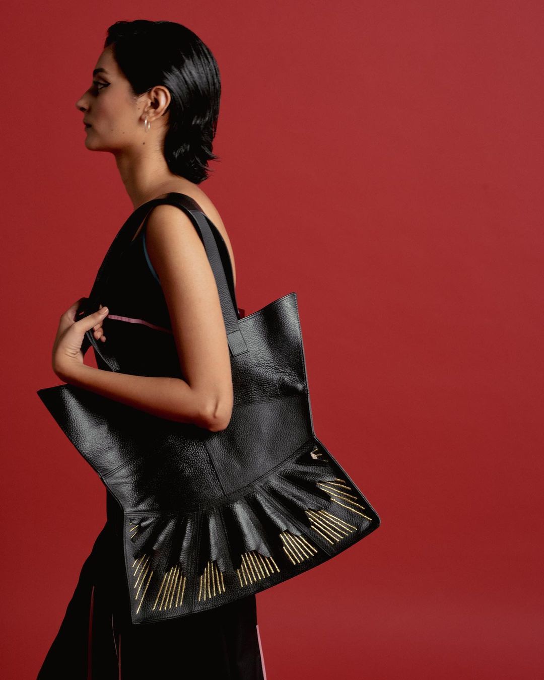 Profile shot of a woman carrying a uniquely designed black leather tote bag with metallic gold fringe accents against a red background.