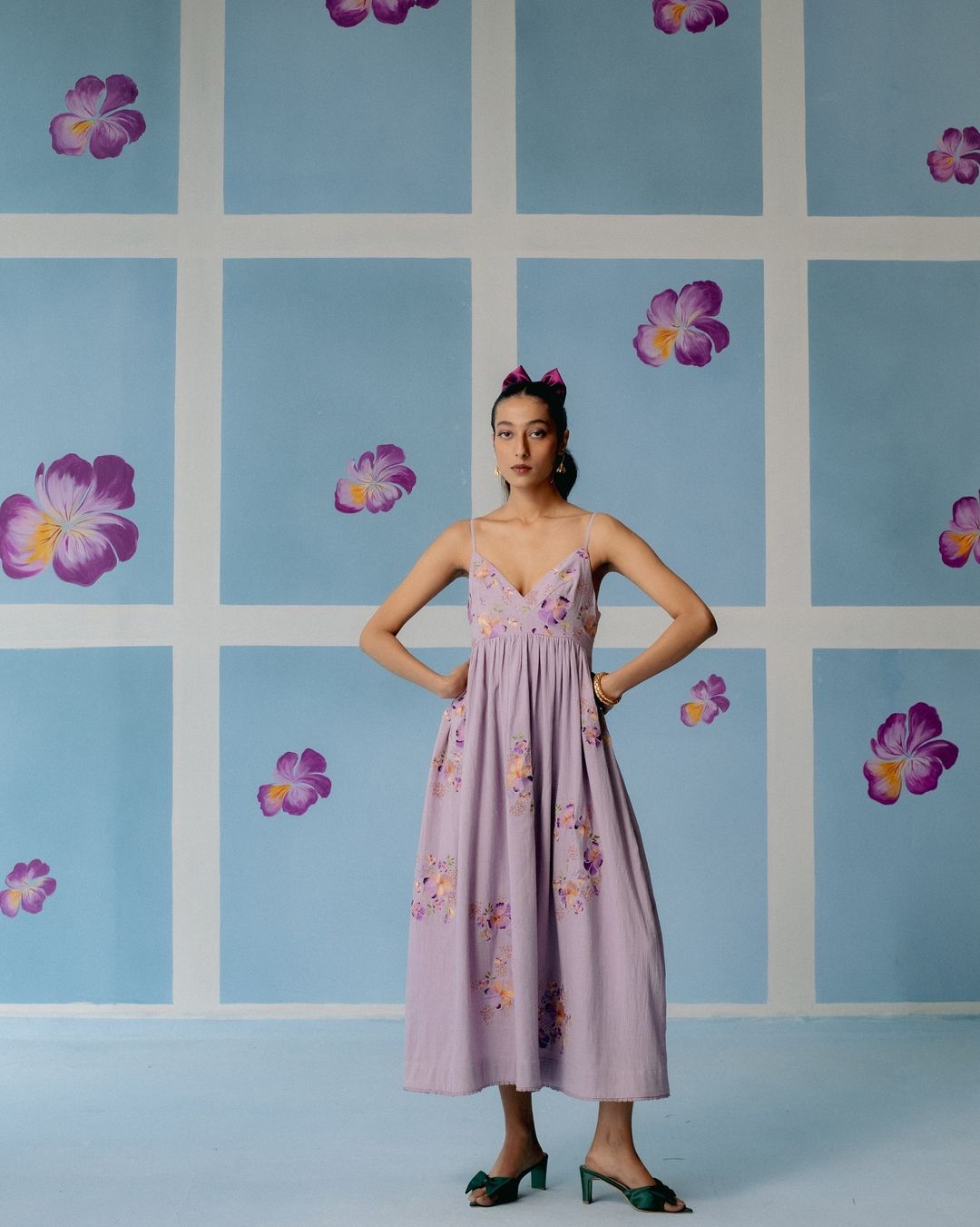 A young woman in a lilac sundress poses in front of a painted blue-and-white floral background, creating a summery, fashionable image.