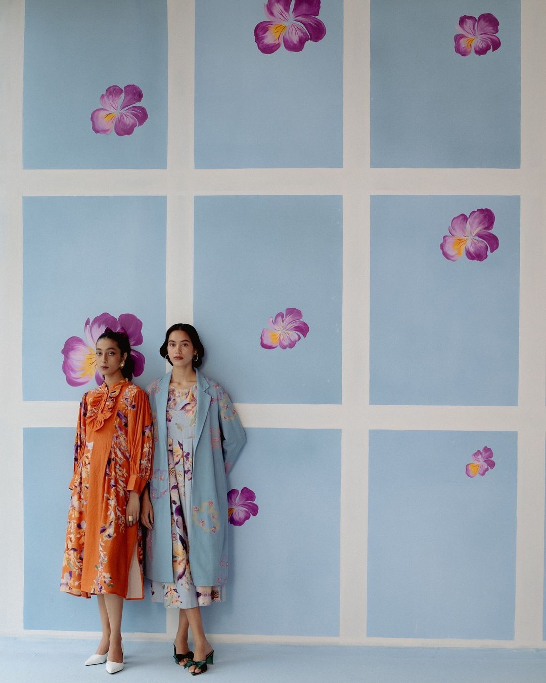 Two women in floral dresses pose in front of a blue wall with painted flowers. Fashion editorial with a colorful, patterned backdrop.