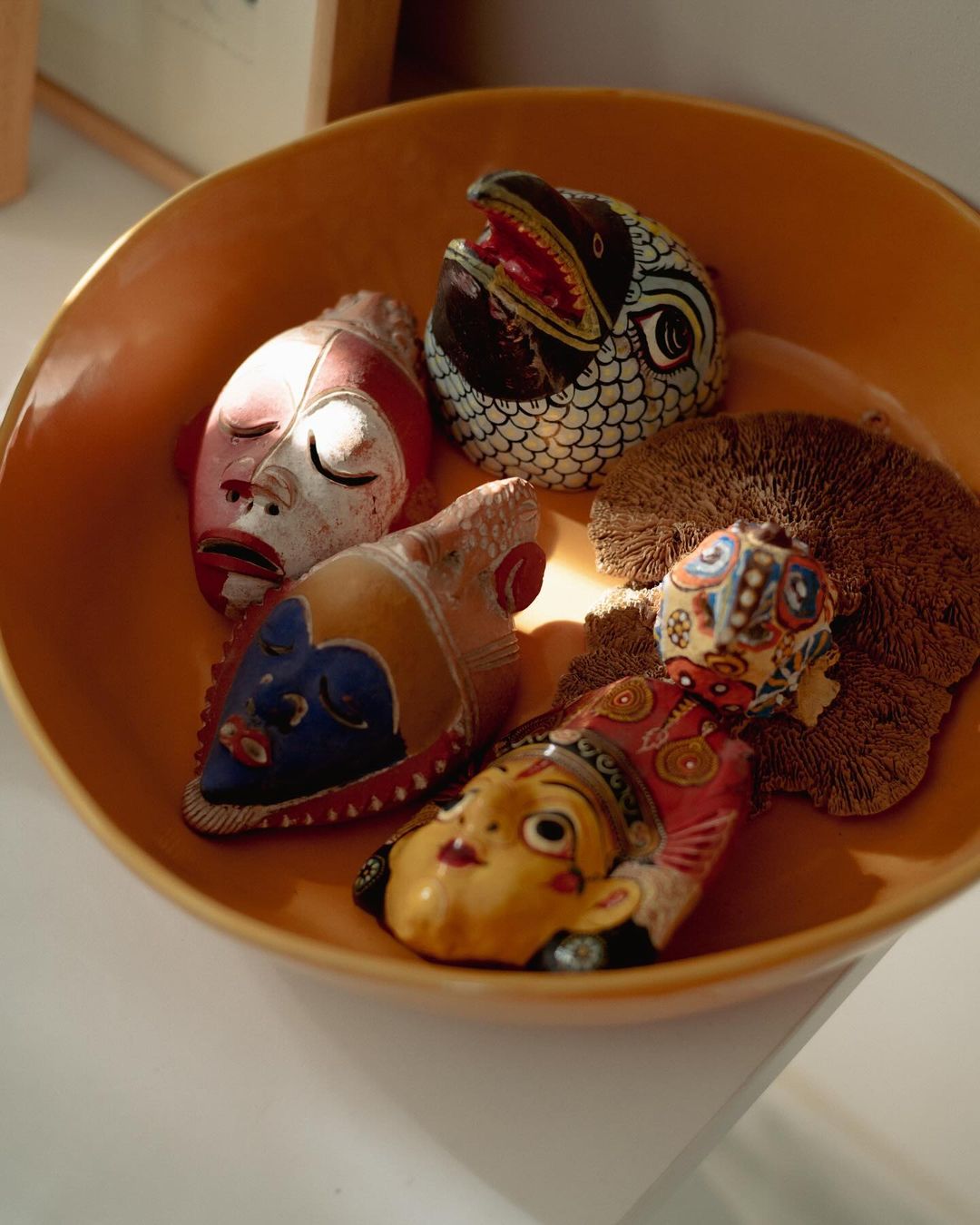A collection of four decorative tribal masks, including a bird-shaped mask, arranged inside a tan ceramic bowl set against a white surface.