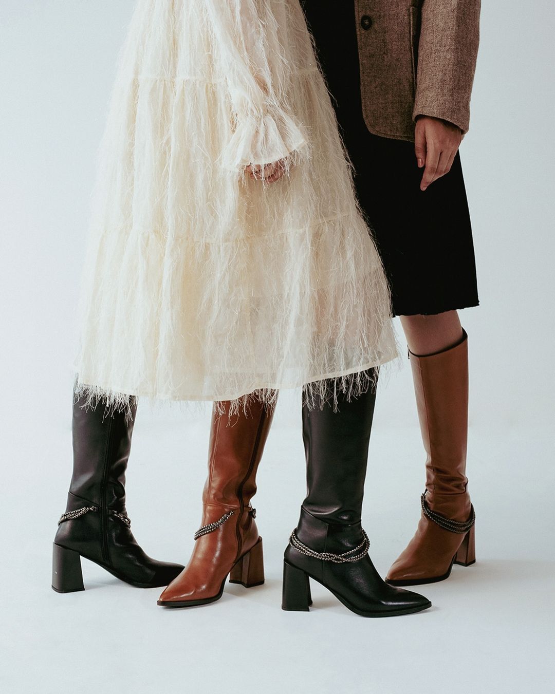Two women showcasing fashionable knee-high boots in brown and black with block heels, paired with a textured white dress and a brown blazer.