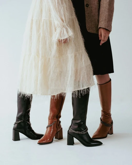 Two women showcasing fashionable knee-high boots in brown and black with block heels, paired with a textured white dress and a brown blazer.