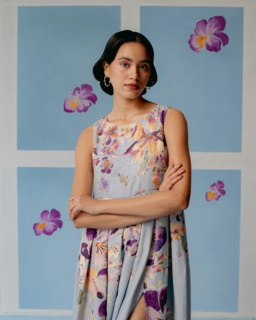 Portrait of a young woman in a floral dress with arms crossed, standing against a blue background with flower paintings.