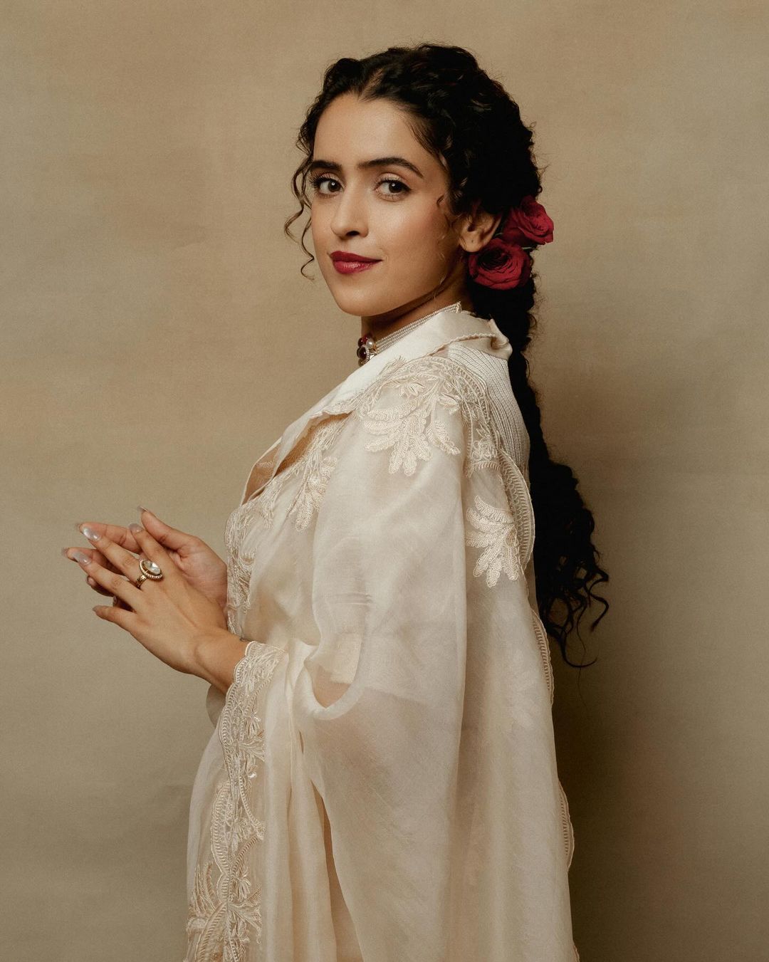 Portrait of a young woman with dark braided hair, wearing a light-colored dress with floral embroidery and red roses in her hair.
