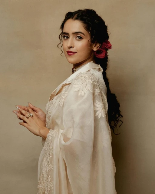 Portrait of a young woman with dark braided hair, wearing a light-colored dress with floral embroidery and red roses in her hair.