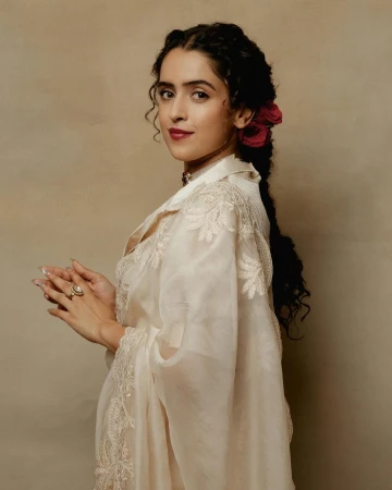 Portrait of a young woman with dark braided hair, wearing a light-colored dress with floral embroidery and red roses in her hair.