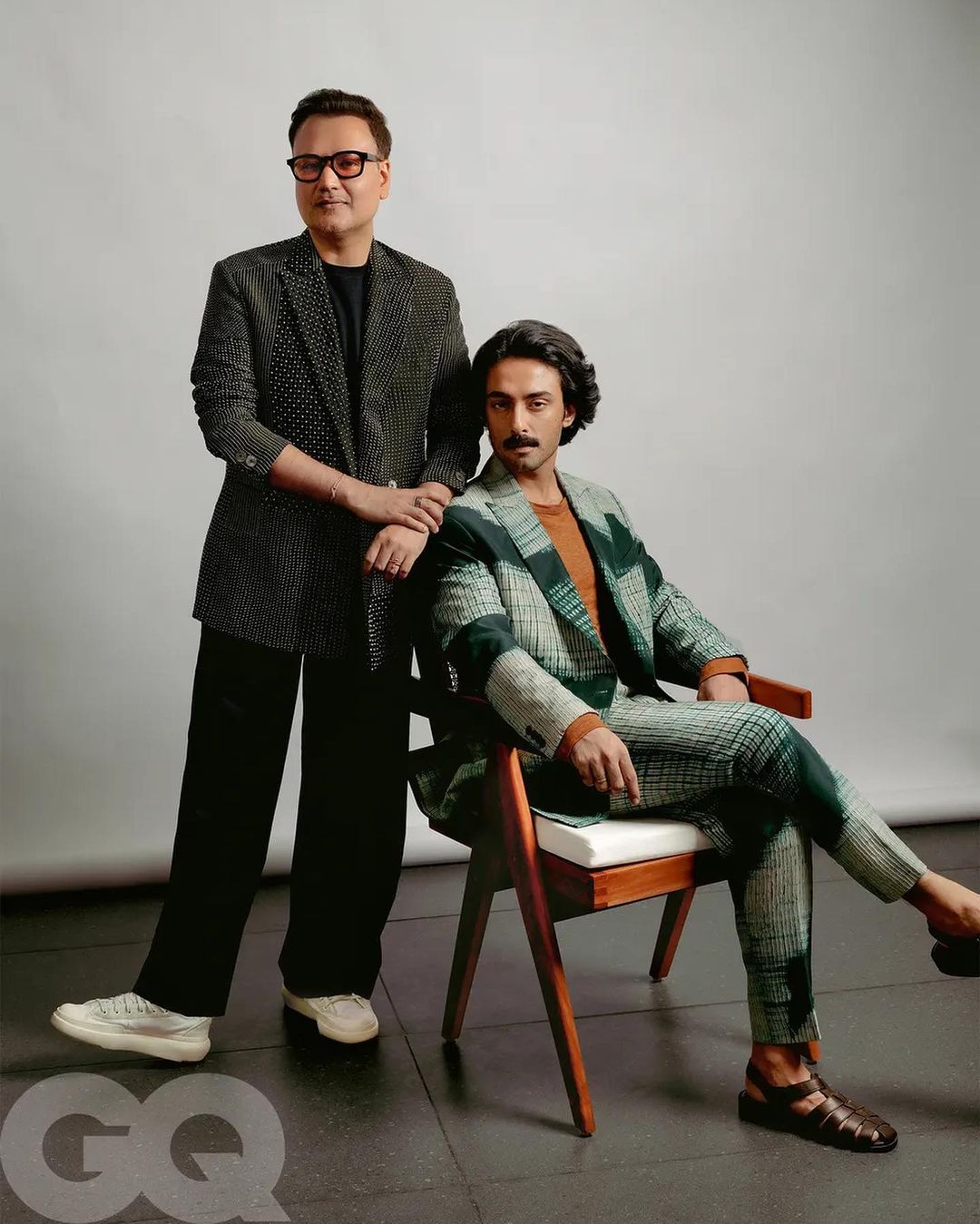 Two men pose in patterned suits against a neutral background. One man sits in a wood chair while the other stands next to him.