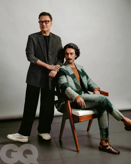 Two men pose in patterned suits against a neutral background. One man sits in a wood chair while the other stands next to him.
