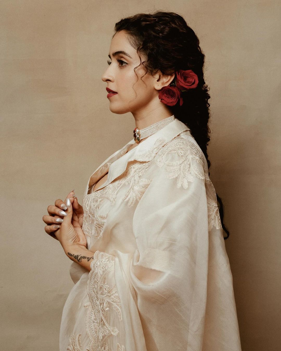 Portrait of a woman with curly hair and red roses, wearing a light-colored, embroidered outfit, and a pearl necklace against a neutral background.