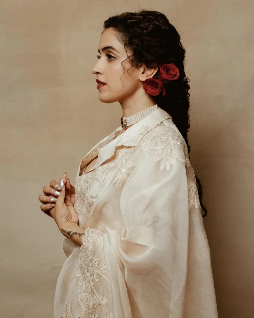 Portrait of a woman with curly hair and red roses, wearing a light-colored, embroidered outfit, and a pearl necklace against a neutral background.