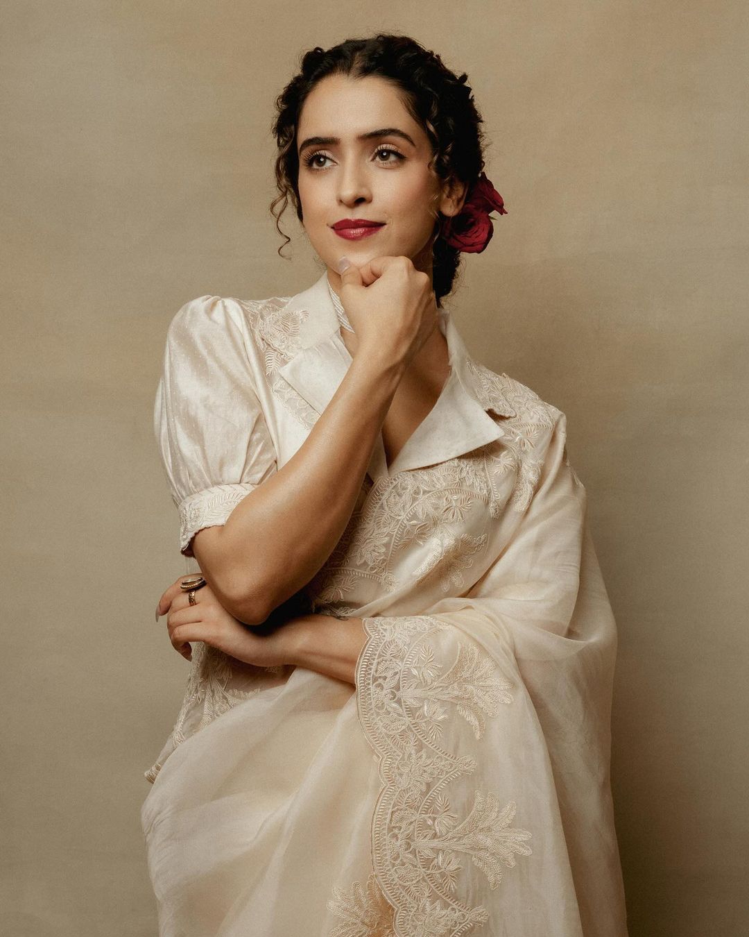 A woman wearing an embroidered formal sari with a flower in her hair, poses in a studio with a neutral background, looking off camera.