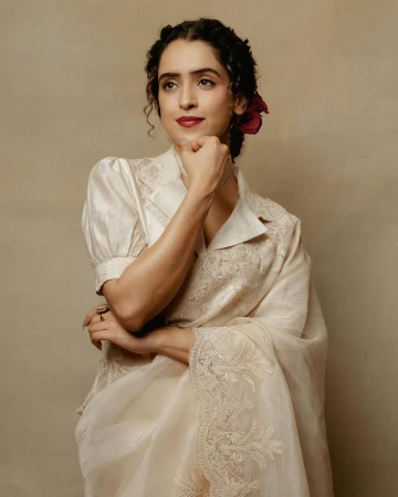 A woman wearing an embroidered formal sari with a flower in her hair, poses in a studio with a neutral background, looking off camera.