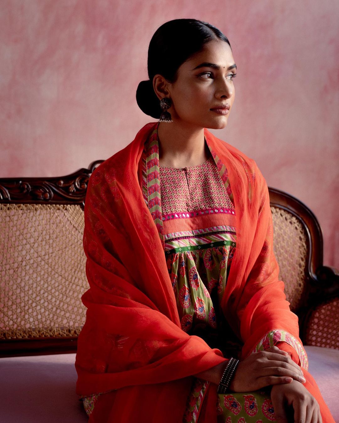 Adult female in a traditional dress and orange shawl, sitting on a decorative chair with a pink textured background, looking to the side.