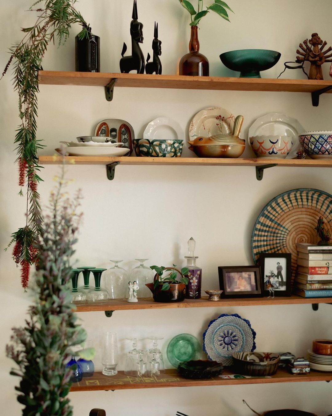 Rustic wooden shelves display diverse pottery, glassware, vases, figurines, framed photos, books, and decorative items against a neutral wall.