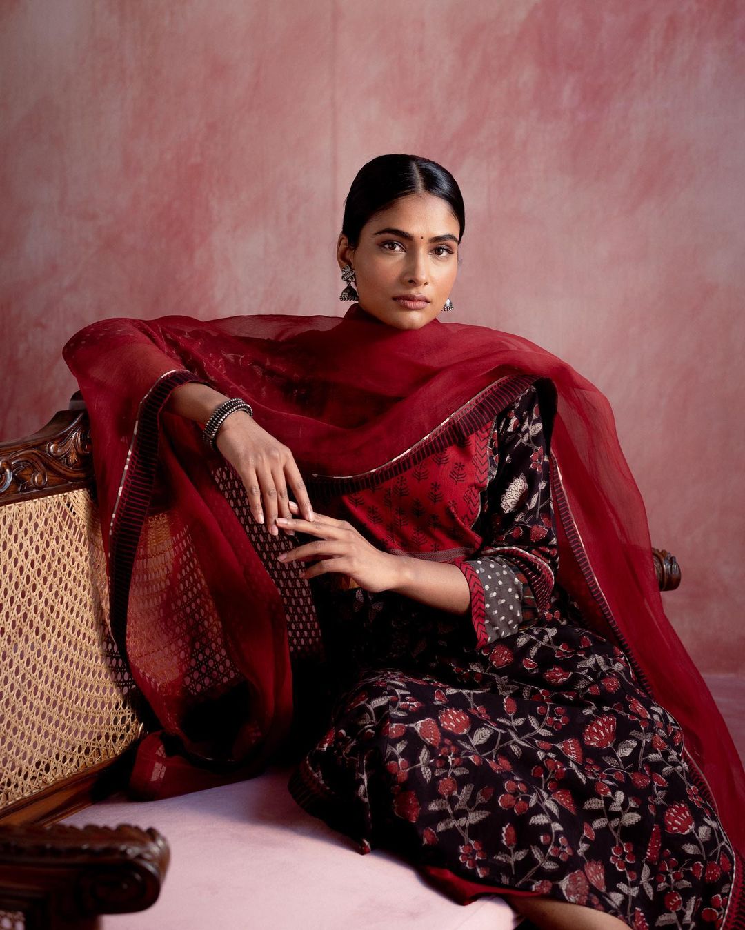 Woman in traditional Indian salwar kameez with floral pattern and dupatta, seated on carved wooden furniture, studio setting.