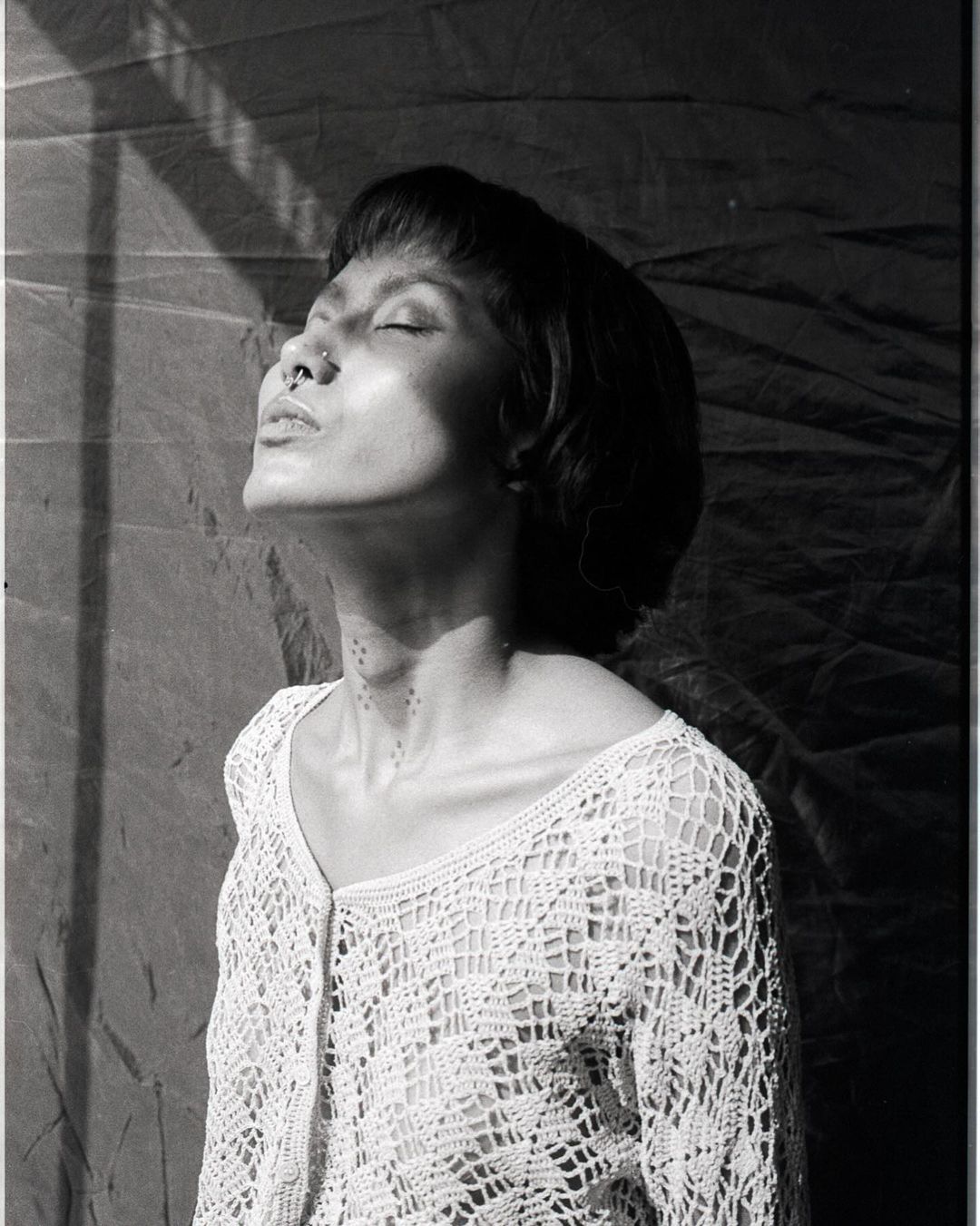 Black and white portrait of a woman with short hair, wearing a crocheted top, her face tilted up, eyes closed, in soft window light.