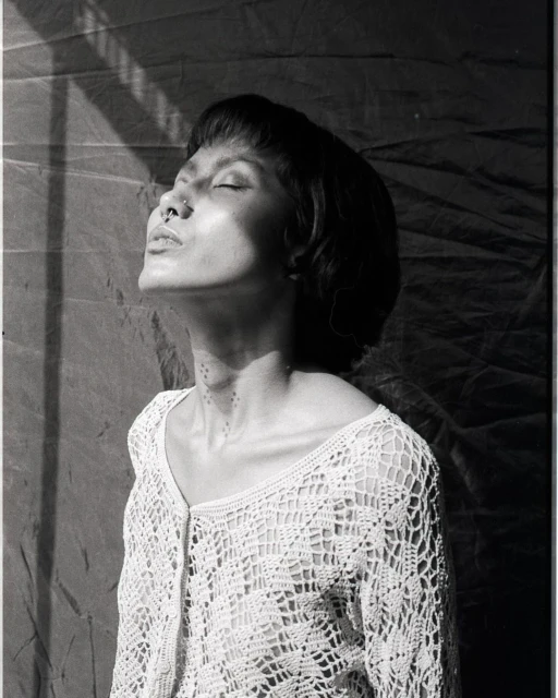 Black and white portrait of a woman with short hair, wearing a crocheted top, her face tilted up, eyes closed, in soft window light.