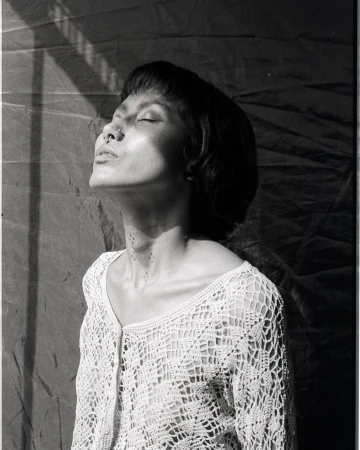 Black and white portrait of a woman with short hair, wearing a crocheted top, her face tilted up, eyes closed, in soft window light.