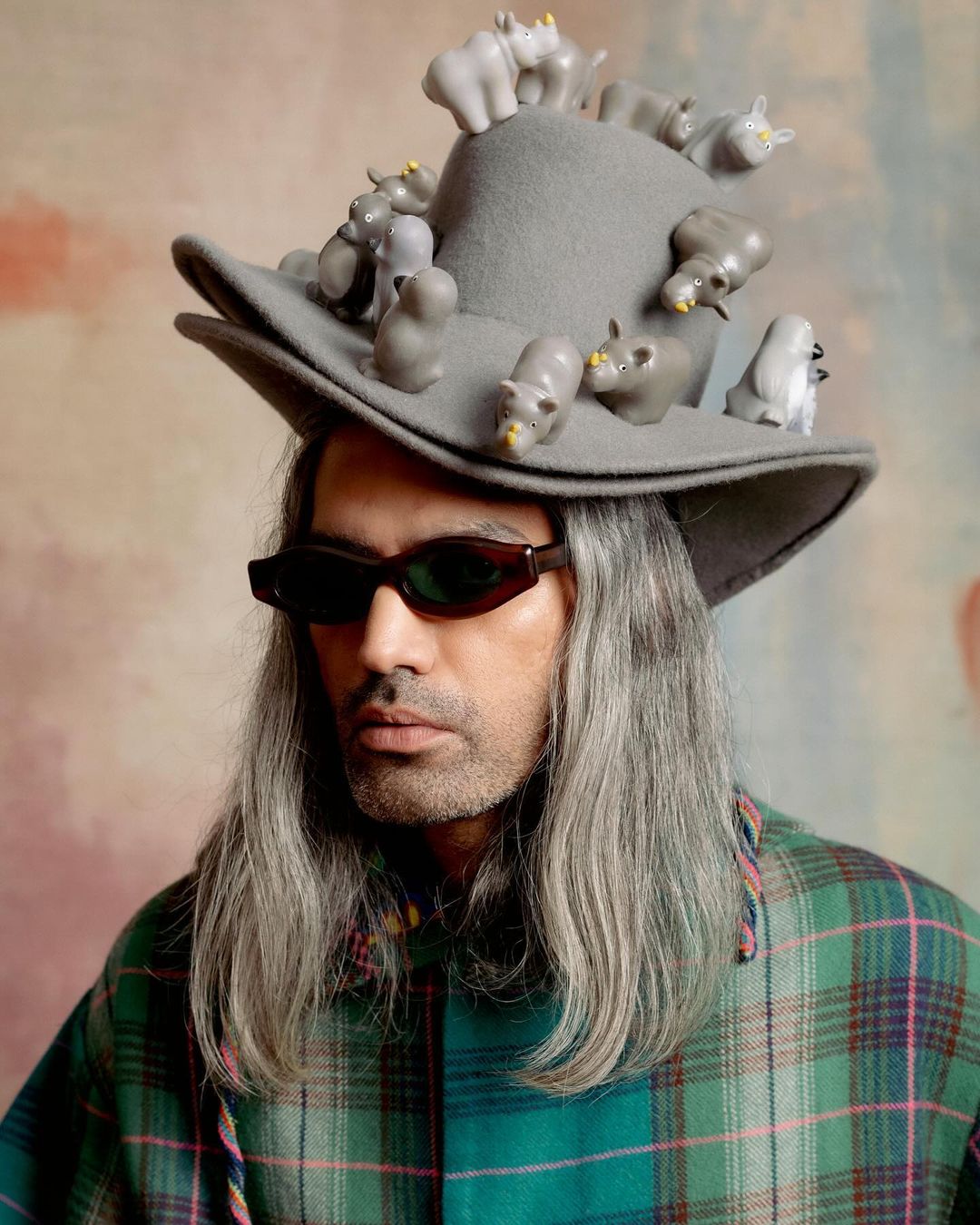 Adult man wearing green plaid shirt, sunglasses, and gray hat decorated with hippo figurines, standing against a colorful backdrop.