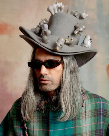 Adult man wearing green plaid shirt, sunglasses, and gray hat decorated with hippo figurines, standing against a colorful backdrop.