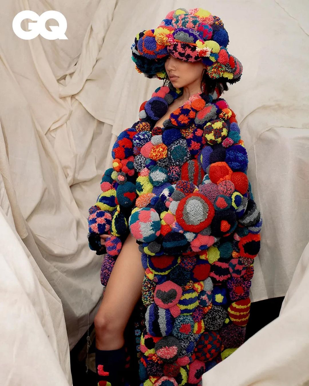 Fashion editorial shot of a woman wearing a multicolored pom-pom coat and matching hat, posing against a textured off-white backdrop indoors.