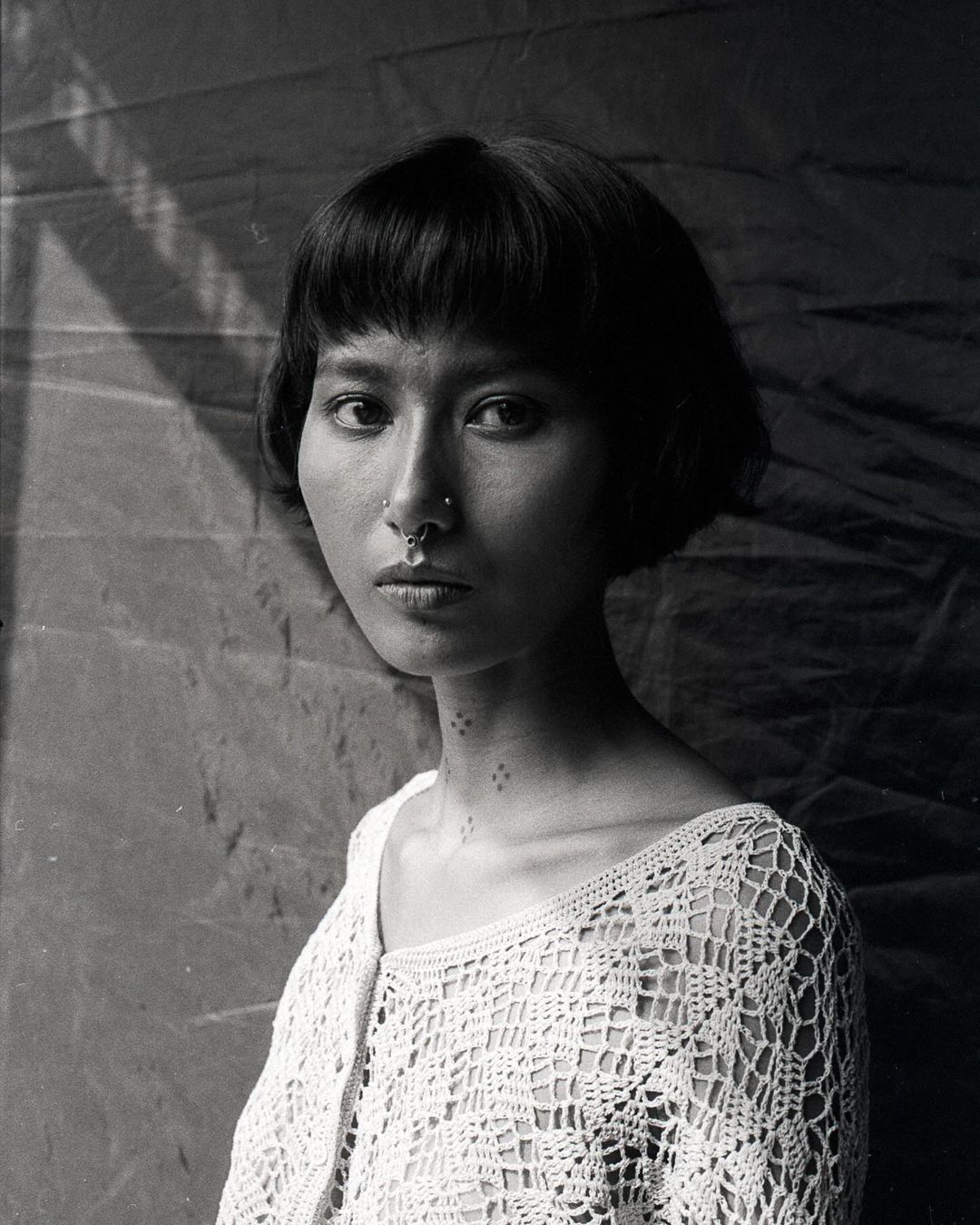 Monochrome portrait of a young woman with a nose ring, wearing a crocheted top, and posing against a softly textured background.