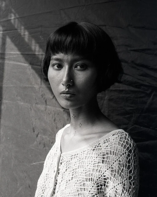 Monochrome portrait of a young woman with a nose ring, wearing a crocheted top, and posing against a softly textured background.
