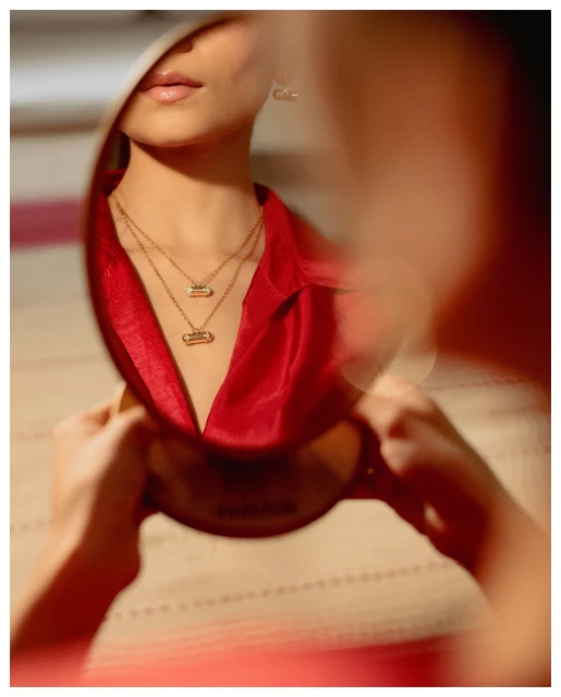 Reflection captures a woman's face, neck, and red silk blouse, accentuated by layered gold pendant necklaces and natural studio lighting.