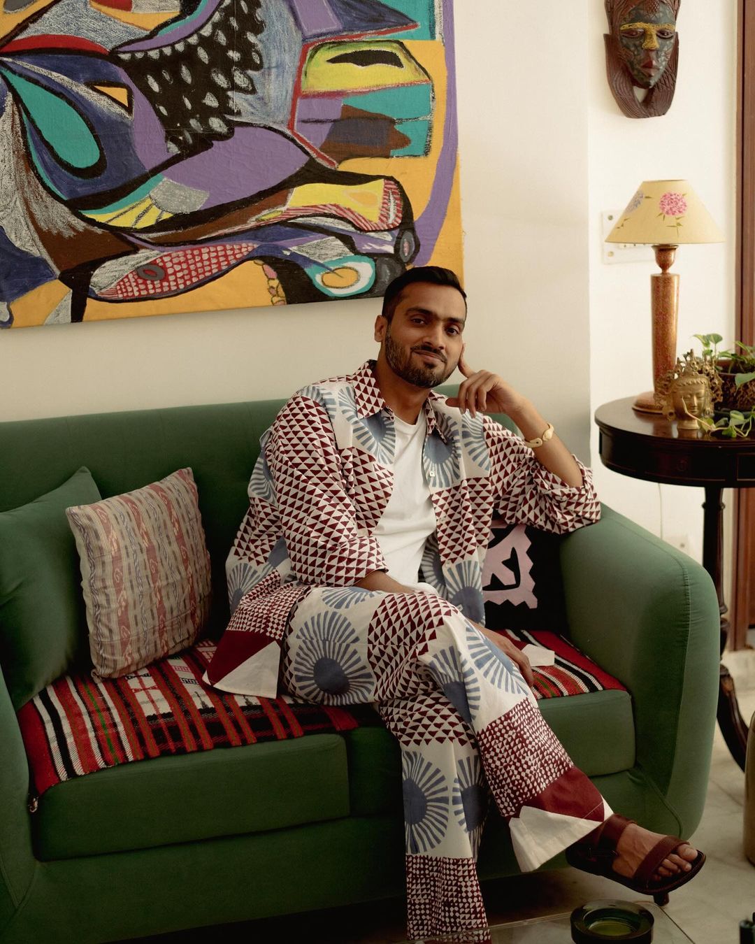 Adult male in patterned shirt and pants sitting on green sofa in living room with colorful abstract art and decor. Indoor setting.