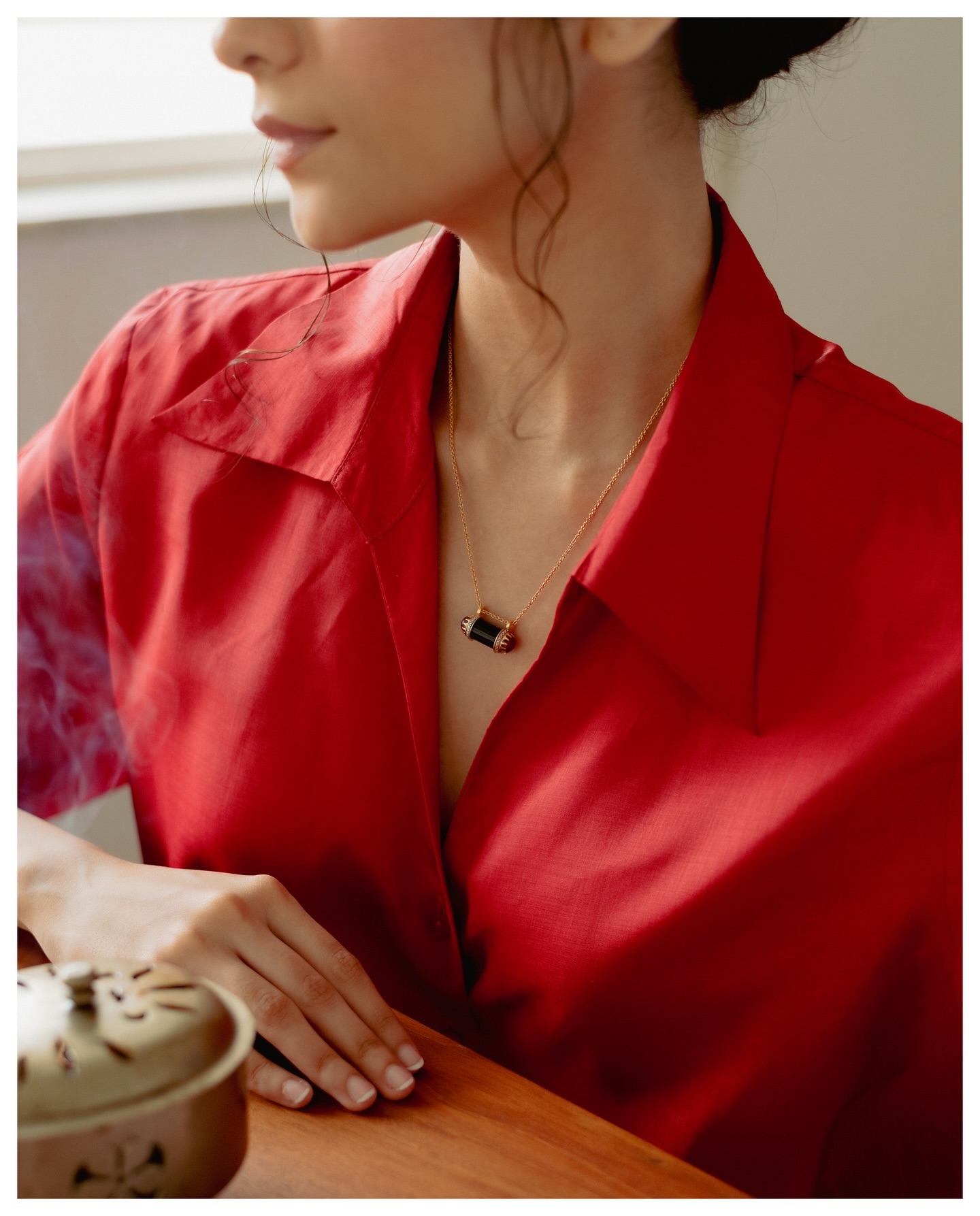 Woman in red shirt poses with gold necklace indoors. Wooden table visible with a ceramic incense burner in soft, warm lighting.