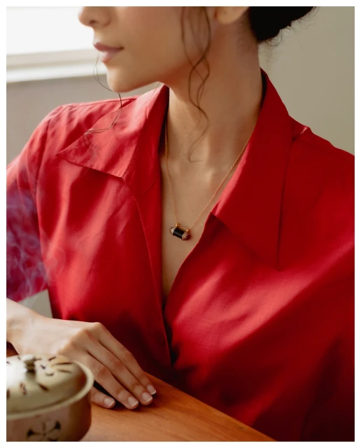 Woman in red shirt poses with gold necklace indoors. Wooden table visible with a ceramic incense burner in soft, warm lighting.