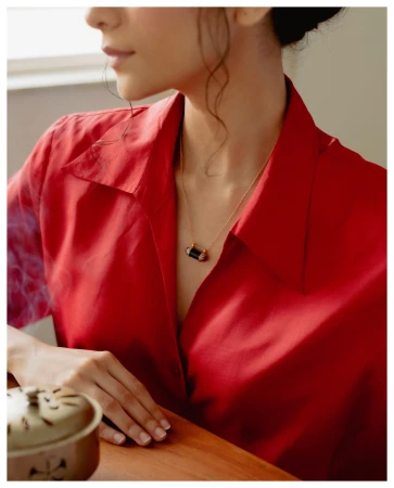 Woman in red shirt poses with gold necklace indoors. Wooden table visible with a ceramic incense burner in soft, warm lighting.